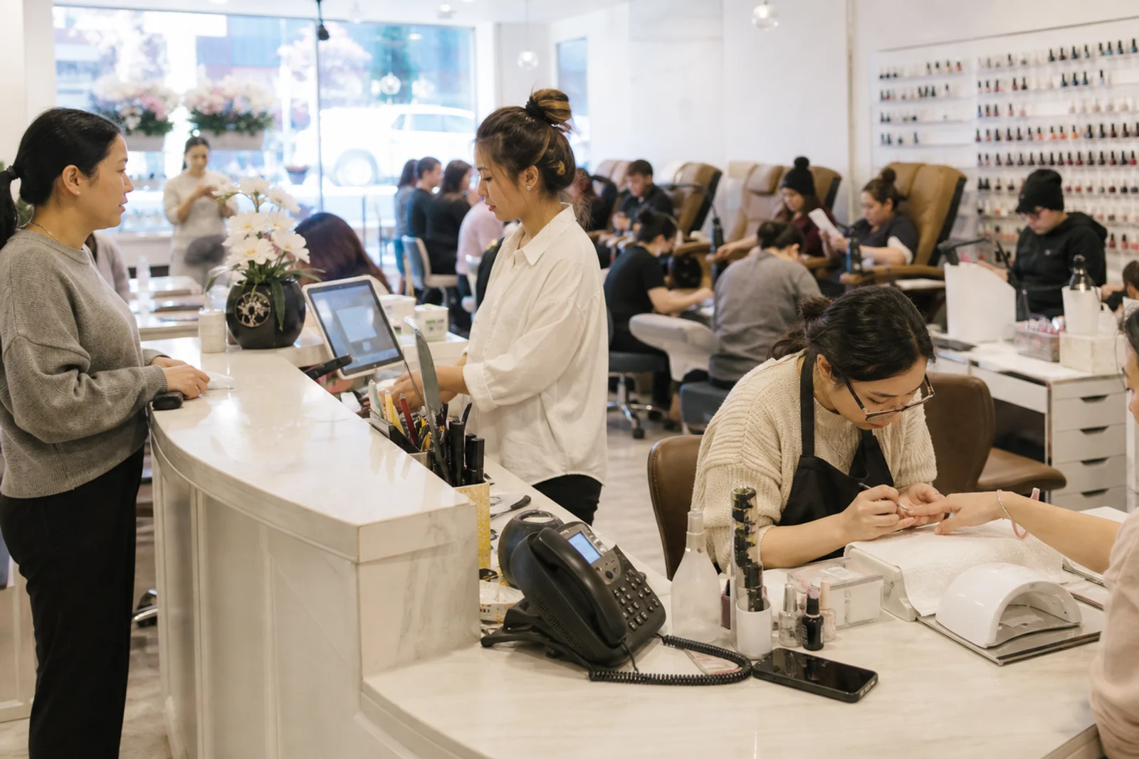 Busy nail salon with technicians serving clients and staff at the front desk